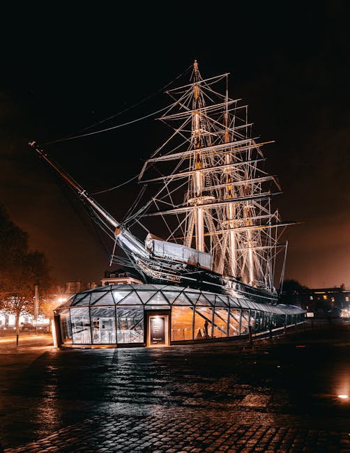 The image depicts a nighttime scene with a large, historic sailing ship displayed on land, illuminated by orange and white lights that highlight its tall masts, rigging, and detailed wooden hull. The 
